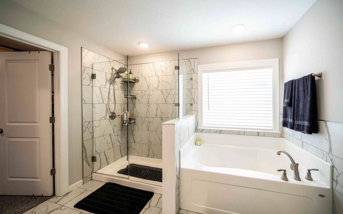 Marble-tiled glass shower next to a deep soaking tub under a window